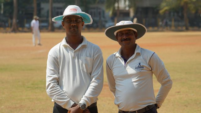 Two cricket umpires stand on a dusty field